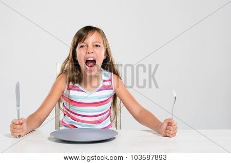 Hungry angry young girl screaming, shouting, throws a tantrum sitting at a table with empty plate