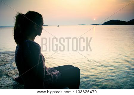 Young Woman Sitting On The Beach At Sunset, A Symbol Of Dreams And Longing