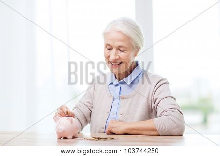 savings, money, annuity insurance, retirement and people concept - smiling senior woman putting coins into piggy bank at home