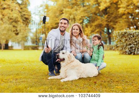 family, pet, season, technology and people concept - happy family with labrador retriever dog taking picture by smartphone on selfie stick in autumnl park