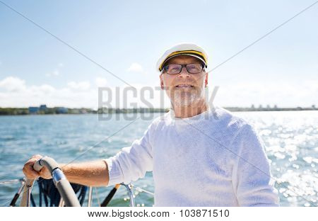 sailing, age, tourism, travel and people concept - happy senior man in captain hat on steering wheel and navigating sail boat or yacht floating in sea