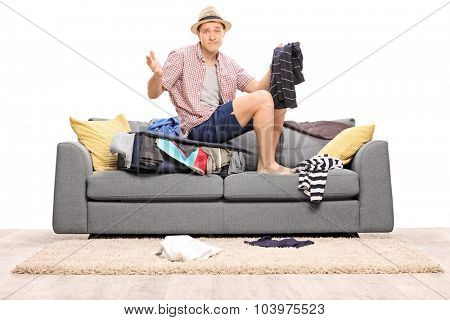 Sad young man sitting on his overstuffed suitcase and gesturing with his hands isolated on white background
