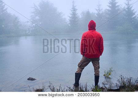 Rear view of man in waterproof jacket holding hands in pocket and looking at lake