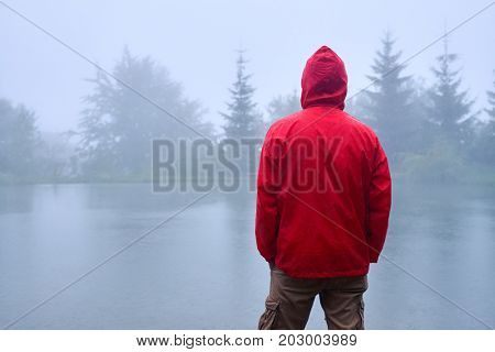 Rear view of man in waterproof jacket standing near lake and holding hands in pocket 