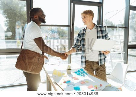 You are hired. Professional young office worker holding his laptop and shaking hand of a new emploee while standing in the office