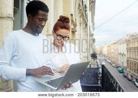 Multi-ethnic team of talented designers working together on joint project while breathing fresh air on balcony, picturesque view of city street