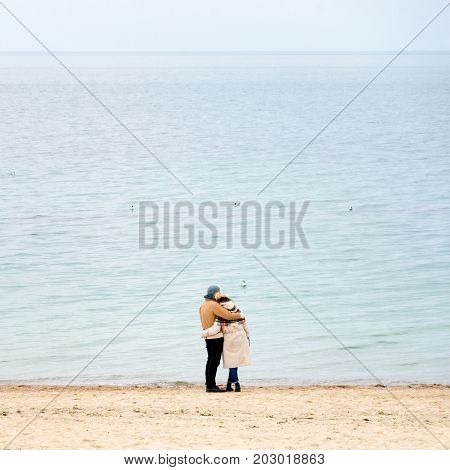 Happy loving couple embracing wearing warm clothes standing on ocean coast. Cold season, sea shore, empty beach in autumn or winter