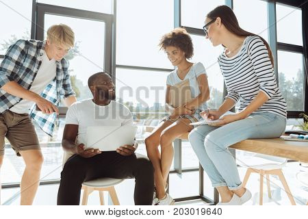Lets do it together. Cheerful afro american male student using his laptop and talking with his groupmates while getting ready with the university project