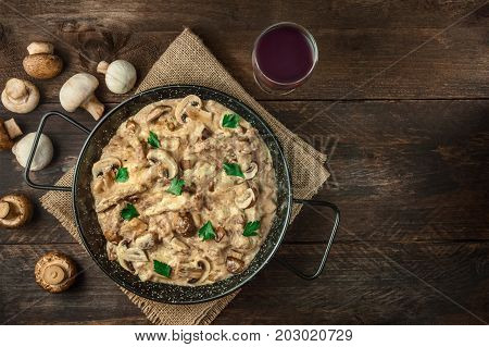 Mushroom beef stroganoff, with cremini and champignons, in a frying pan, with a glass of red wine, shot from above on a dark rustic texture with a place for text