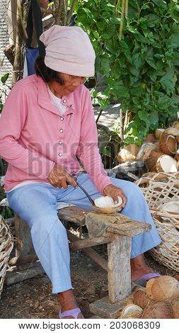 DAVAO ORIENTAL, PHILIPPINES--MARCH 2016: A woman shells coconut meat with a sharp knife outdoors while sitting on a wooden bench.