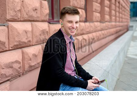 Cheerful happy young man in plaid shirt and jacket listening to music with headphones from cell phone and singing outdoors. Student with smart phone and headphones