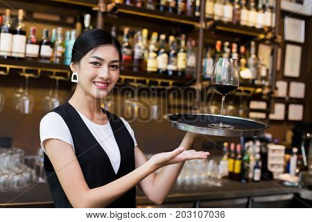 Young Asian Woman Waiter Restaurant Catering Service Happy Emotion. Woman Waiter Present Red-Wine for Customer at Bar. Woman with Wine in Bar Concept.