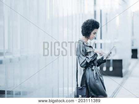 Charming young afro american business girl is standing outdoor of contemporary office near modern regular glass striped construction and using her digital tablet with copy space for your logo or text