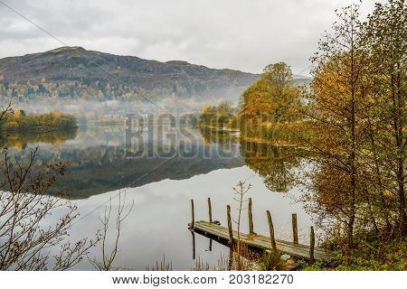 Still water on Grasmere with reflections of Autumn colours and a small landing jetty in the foreground