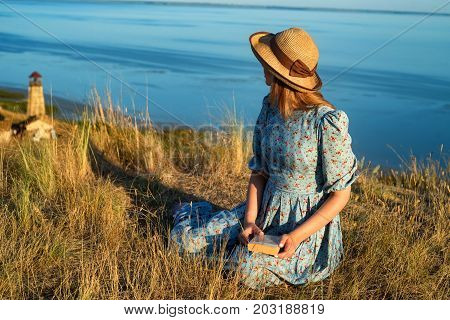 Pretty vintage girl wearing dress and hat sitting alone on hill at seaside fantastic background