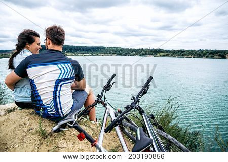 couple sits and looking at the lake two bicycles near them zure water on background