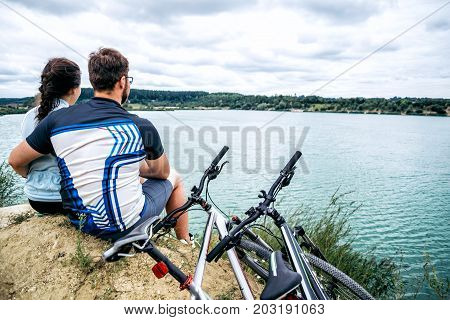 couple sits and looking at the lake two bicycles near them zure water on background
