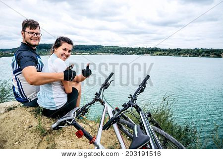 cuple of sportmen sitting near lake with bicycles and showing thumb up