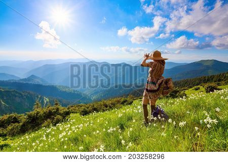 On the lawn in mountains landscapes the hipster girl in dress stockings and straw hat stays watching the sky with clouds.