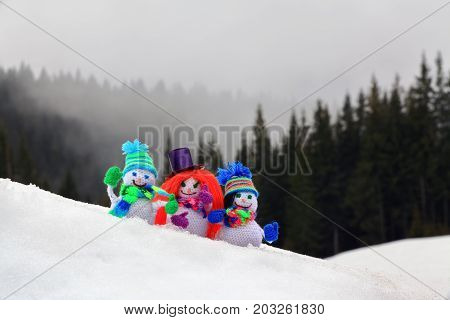 Three funny small knitted with a crochet hook snowmen dressed in colorful hats scarves and gloves standing in snow against a background of misty spruce forest high in the mountains.