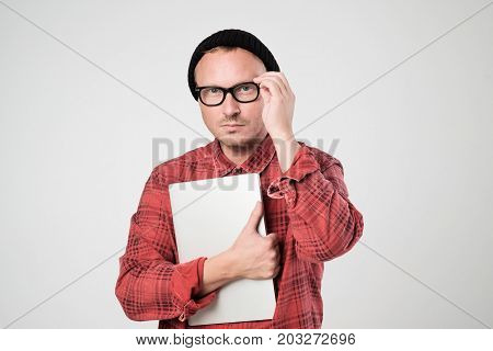 A young programmer in a black hat is holding a laptop in his hands. He straightens his glasses and looks doubtfully into the camera. Concept oh negative human emotion