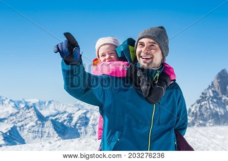 Young father enjoying with daughter a sunny winter day. Happy father with daughter enjoying winter vacation over mountains. Cheerful father giving piggyback ride to daughter while pointing mountain.