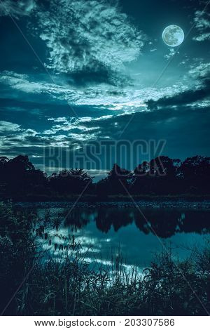Sky With Dark Cloudy And Full Moon Above Silhouettes Of Trees And Tranquil Lake.