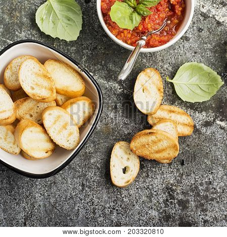 Healthy snack or lunch. Homemade tomato vegetable sauce with garlic, herbs, salt, butter, spices, fresh basil in a ceramic bowl with grilled toasts on a gray stone background. Top View