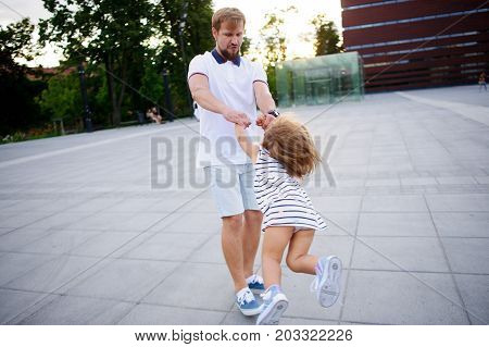 Young dad is playing with a charming little daughter. The man holds the baby by hands and is turned together with her. The child rejoices.