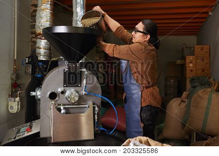 Roastery owner putting container of green coffee beans into roaster