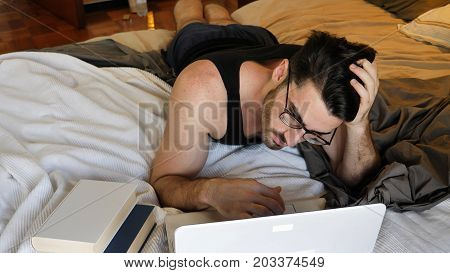 Attractive Young Man with Serious Expression, with Laptop on Bed Working on his Start-up Business, with Books Next to Him - Young Male College or University Student Doing Homework, in Bedroom