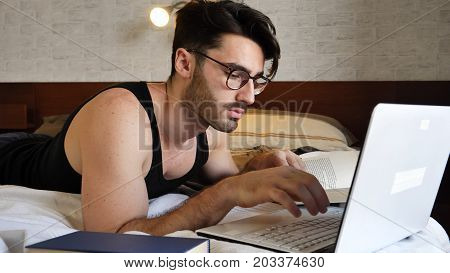 Attractive Young Man with Serious Expression, with Laptop on Bed Working on his Start-up Business, with Books Next to Him - Young Male College or University Student Doing Homework, in Bedroom