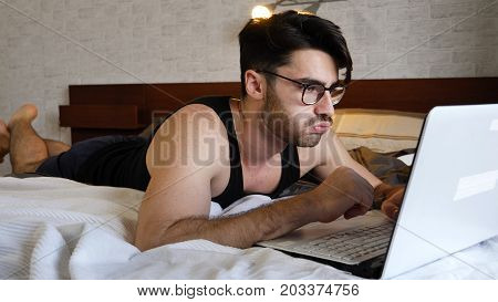 Tired, Fed-Up Young Man with Serious Expression, with Laptop on Bed Working on his Start-up Business - Young Male College or University Student Doing Homework, in Bedroom