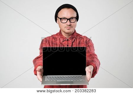 A young programmer in a black hat is holding a laptop in his hands. He shows screen of notebook. Concept oh negative human emotion