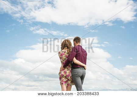 View from the back of a young couple stands in an embrace and looks out into the distance against the sky with clouds. A vision for the future