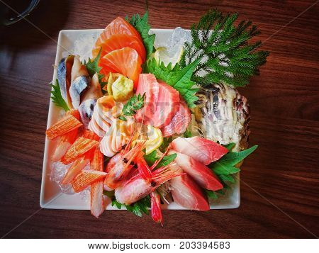 Mixed sashimi set on white plate put on wood background. Japanese food style with raw fishes, shrimp, surimi (imitation crab meat) keeping fresh on ice decorated with green vegetables - top view
