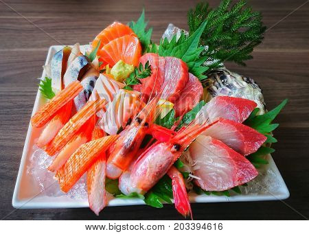 Mixed sashimi set on white plate put on wood background. Japanese food style with raw fishes, shrimp, surimi (imitation crab meat), wasabi; keeping fresh on ice decorated with green leaves