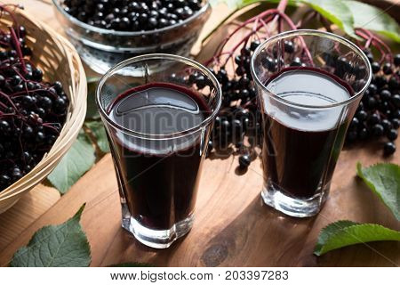 Two glasses of elderberry syrup on a wooden table with elderberries in the background