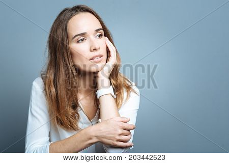 Too many thoughts. Cheerless nice thoughtful woman holding her cheek and thinking about her life while sitting against grey background