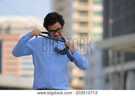 Portrait of stressed young Asian business man removing his tie on urban building in the city background.