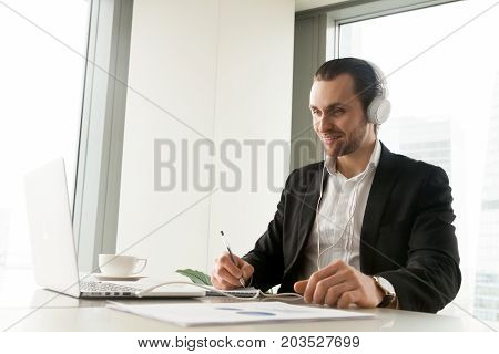 Smiling businessman in headphones taking notes in front of laptop at workplace. Friendly young manager participating in online meeting or conference, remote job interview, learning foreign languages.