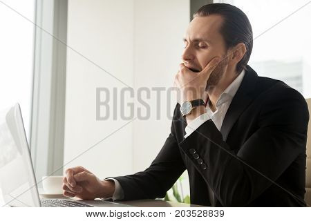 Tired sleepy young businessman yawning in front of laptop at workplace. Project manager exhausted during work hours, CEO struggling staying in focus, entrepreneur feeling fatigue and being drowsy.