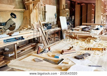 Interior of spacious messy workshop: table with wooden planks, woodworking tools and sawdust on foreground, no people