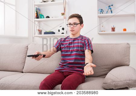 Teenager boy watching television, using remote control to switch channels. Youngster unhappy at what he sees on TV screen. Sitting on couch in living room at home