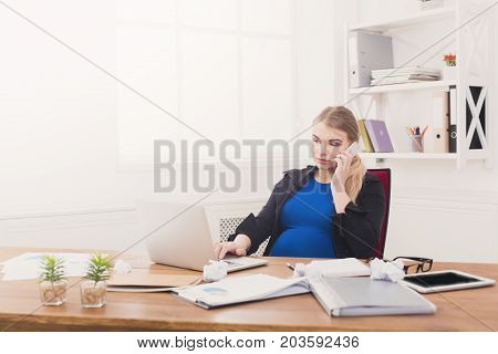 Pregnant business lady at work talking on phone. Worried expectant businesswoman sitting at her working place in office, copy space