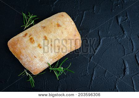 Freshly baked loaf of traditional italian bread ciabatta on black stone table with rosemary. Top view. Copy space.