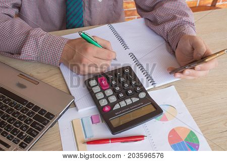 Man hand using calculator and writing make note with calculate about cost at home office. The Man and computer are using a calculator on the table in the office room