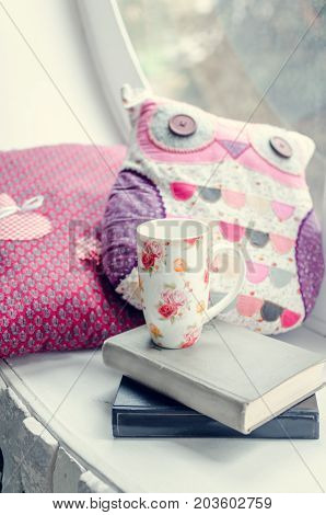 Warm and cozy window seat with cup of coffee or tea with books and cushions. Provence style rustic home decor. Lifestyle background in pink and purple tones.