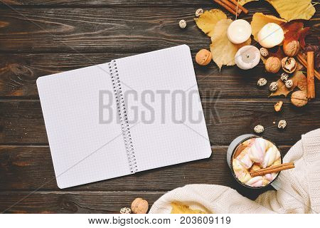Autumn frame made of dried fall leaves, mug of cocoa with marshmellows, nuts, cinnamon, plaid, apples and open copybook Top view on brown wood background. flat lay, copy space