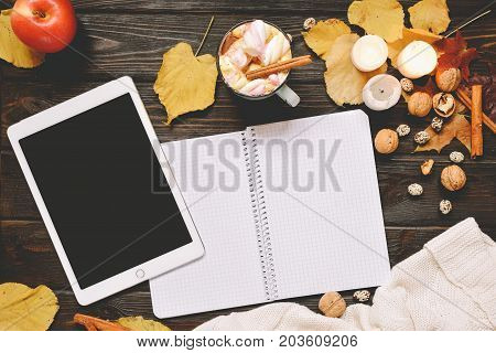 Autumn frame made of dried fall leaves, mug of cocoa with marshmellows, nuts, cinnamon, plaid, apples with tablet mock up. Top view on brown wood background. flat lay, copy space